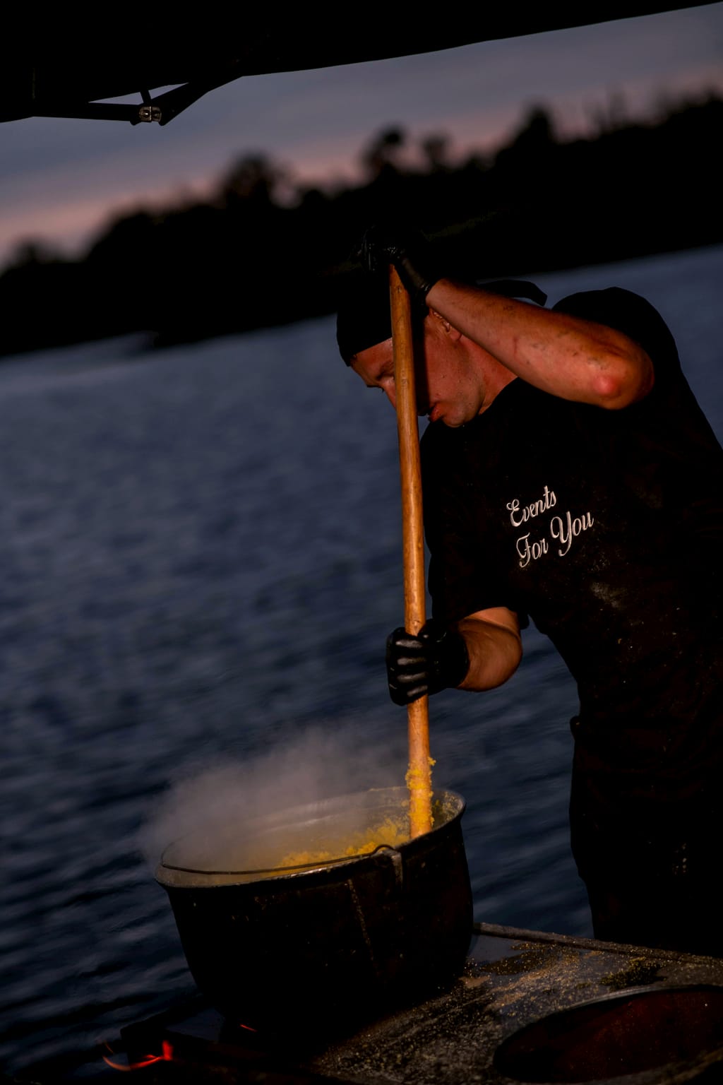 Bucătar mestecând mămăliga la ceaun, la apus, lângă lac